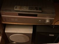 Sony main surround sound unit with remote on top, Polk Audio black subwoofer with round speaker grill, and a black rectangular Polk Audio speaker in the background.