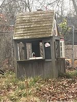 Full view of outdoor wooden toddler playhouse showing pitched shingled roof with moss, open serving window with sink inside, and side arched windows.