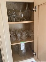 Photo of cabinet shelf with multiple clear glass pitchers, bowls, and several mini cut glass Waterford-style flower vases.