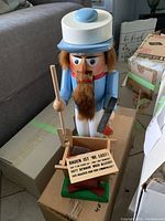Fisherman Nutcracker in blue suit holding a wooden tool and a sign with German script, standing on a green base in front of a sofa and boxes
