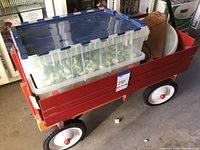 Radio Flyer red wagon with four white and red wheels, containing multiple clear plastic storage containers with blue lids filled with packing peanuts and a bag inside the wagon