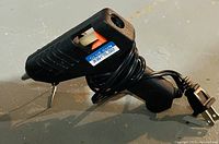 Photo showing black electric hot glue gun with power cord, silver metal stand, and a label indicating 36W 110-220V.