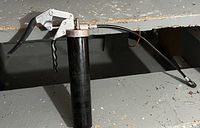 Manual grease gun with a metal lever handle, black cylindrical body, and flexible nozzle tube, photographed on a gray floor beneath a metal workbench.