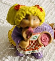 Close-up of one Cabbage Patch Kid figurine with blonde curly hair, purple dress, apron with red checkered pattern, holding candy lollipops.