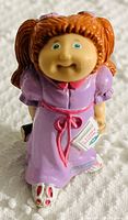 Front view of one PVC Cabbage Patch Kids figurine wearing a purple dress with red ribbon and holding a small book.