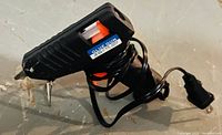 Photo shows a black electric glue gun with an orange trigger and a power cord wrapped around the handle. The gun has a sticker labeling it as a glue gun.