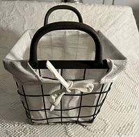 Front angle view of a rectangular metal wire basket with black finish and an off-white removable cloth liner tied in bow knots.