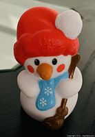 Close-up photo of the snowman little people figure toy showing red hat with pom-pom, blue scarf, orange carrot nose, and broom accessory.