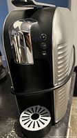 Side view of Verismo K-tee coffee machine showing chrome lever, black front section and circular drip tray.