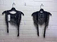 Front view of two black beaded lace shawls side by side on hangers against a brick wall, showing the intricate beadwork and lace patterns.