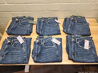 Six pairs of folded new women's jeans on a wooden table, showing a variety of denim washes and tags attached.