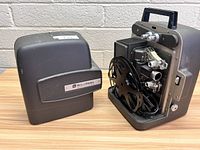 Photo showing projector with attached film reels and power cord, beside its removable protective cover with Bell & Howell Autoload branding.