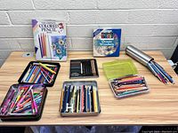 Overview of entire lot showing colored pencils in multiple containers, a metal tin, and two boxed colored pencil books on a table against a brick wall.