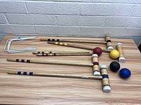 Four wooden croquet mallets with colored stripes, four matching wooden balls in red, yellow, black, blue, and six white-coated metal wickets laid on a wooden surface.