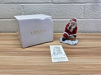 Photo of the Lenox branded white box, Chimney Santa figurine placed next to it, and a folded collector's booklet on a wooden surface against a brick wall background.