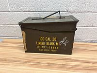 Front side view of olive drab metal ammo box with yellow stenciled text and carrying handle on top.