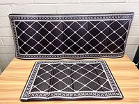 Two black anti-fatigue mats with white diamond and cross pattern, one laid flat and one standing upright against a brick wall, showing the overall design and size.