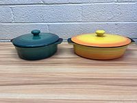 Two oval cast iron casseroles with lids, one green and one yellow-orange gradient, side by side on wooden surface