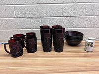 Photo displaying six deep purple glass cups with handles, five matching bowls, one tall vase, and two white ceramic cups with berry and leaf patterns on the right side.