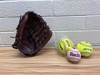 Wilson A800 baseball glove alongside two wrapped yellow Wilson softballs and one white baseball with D-Backs branding placed on a wooden surface against a gray brick wall.