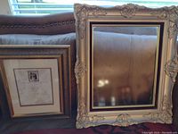 Two picture frames side by side on a couch, the smaller one is simple and dark-toned, the larger one is ornate cream-colored with detailed corners and inner borders.