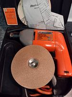 Black & Decker electric drill with attached sanding disc, inside a black hard case showing instruction manual and documents.