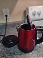 Red Hamilton Beach electric kettle with open hinged lid showing interior and power base detached.
