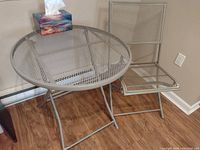 Side view of assembled metal mesh round table with a tissue box on top and matching folding chair beside it, placed on wood floor in corner of room.