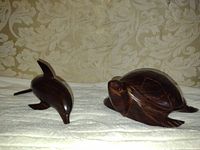 Two hand carved hardwood figurines, a dolphin and a turtle, displayed on white towel against floral wallpaper.