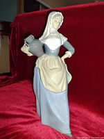 Front angle of the porcelain figurine showing the lady holding a water jug, dressed in blue dress and beige apron with white headscarf.