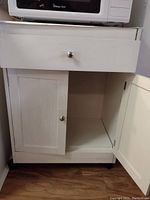 Microwave placed on top of a white wooden cabinet with drawer and double doors, both doors open showing interior storage space and caster wheels visible at bottom