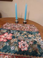 Photo shows four floral cloth placemats with padded dark green backing and two silver candle holders with blue taper candles on a wooden table.