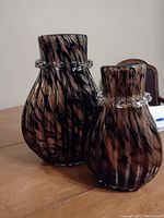 Two glass vases with gold and black tiger stripe pattern and clear knotted glass bands around the necks arranged on a wooden table.