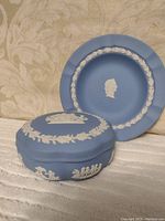 Blue Wedgwood Jasperware covered dish and matching plate displayed side by side, showing raised white classical relief motifs on matte blue background.