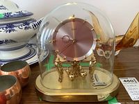 Front view of Kundo anniversary clock with glass dome covering brass base and pink dial. Winding key placed inside dome.