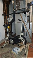 Full left side view of CCM exercise bike in basement showing frame, seat, and part of the flywheel guard with broom and horseshoes partially visible in the background.
