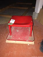Red metal mechanics cart with a square top and bottom platform, mounted on casters, placed on an orange floor.