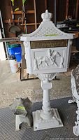 Front view of white cast iron pedestal mailbox with 'LETTERS' panel featuring horse and rider relief and separate grey decorative cap in foreground
