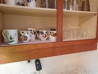 Shelf displays five fruit-themed ceramic mugs with handles and six clear drinking glasses with diamond pattern, both sets on shelf with white lace liner.
