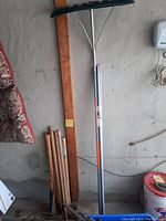 Photo showing wooden handled axes, hammers and sledge hammer leaning against wall near two extension poles.