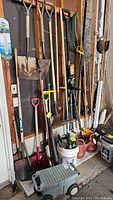 Overview of long handled gardening tools hung on wall and standing, with watering cans and bucket below