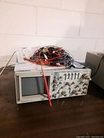 Front view of Kenwood CS-5170 oscilloscope with CRT display and red probe attached, pile of cables on top.