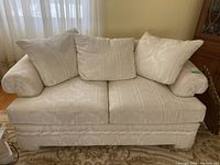 Front view of cream-colored upholstered love seat with three back cushions and two seat cushions on patterned rug near window with sheer curtains.
