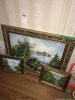 Three framed canvas paintings leaning against wall on wooden floor, with ornate gold/bronze frames. Different sizes shown.