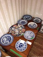Wide view of 10 decorative plates on two wooden surface areas arranged to show variety including blue and white plates and large colorful Imari Ware plate.