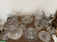 Photo showing a variety of pressed glass items including a large bowl, sugar bowl, creamer, and smaller bowls and plates on a wooden surface.