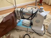 Side view of the sewing machine with brown cover, power cord, and small container on base.