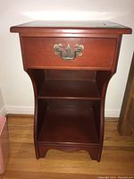 Front view of one wood side table showing drawer with ornate metal pull and open shelf below.