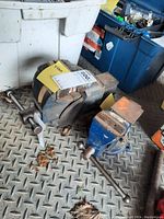 Two metal workbench vices displayed on a diamond plate metal floor, showing the larger vice at the back and smaller vice in front with their handles, jaws and bases visible.