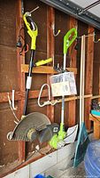 Two electric weed trimmers and one plastic fan rake hanging on garage wall. Ryobi trimmer is yellow with black shaft; Greenworks trimmer is green and silver. Blue plastic fan rake below trimmers.